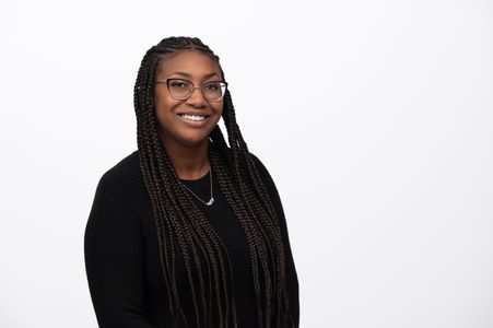 A Black woman with long braids, glasses, and a black sweater smiles against a white background.
