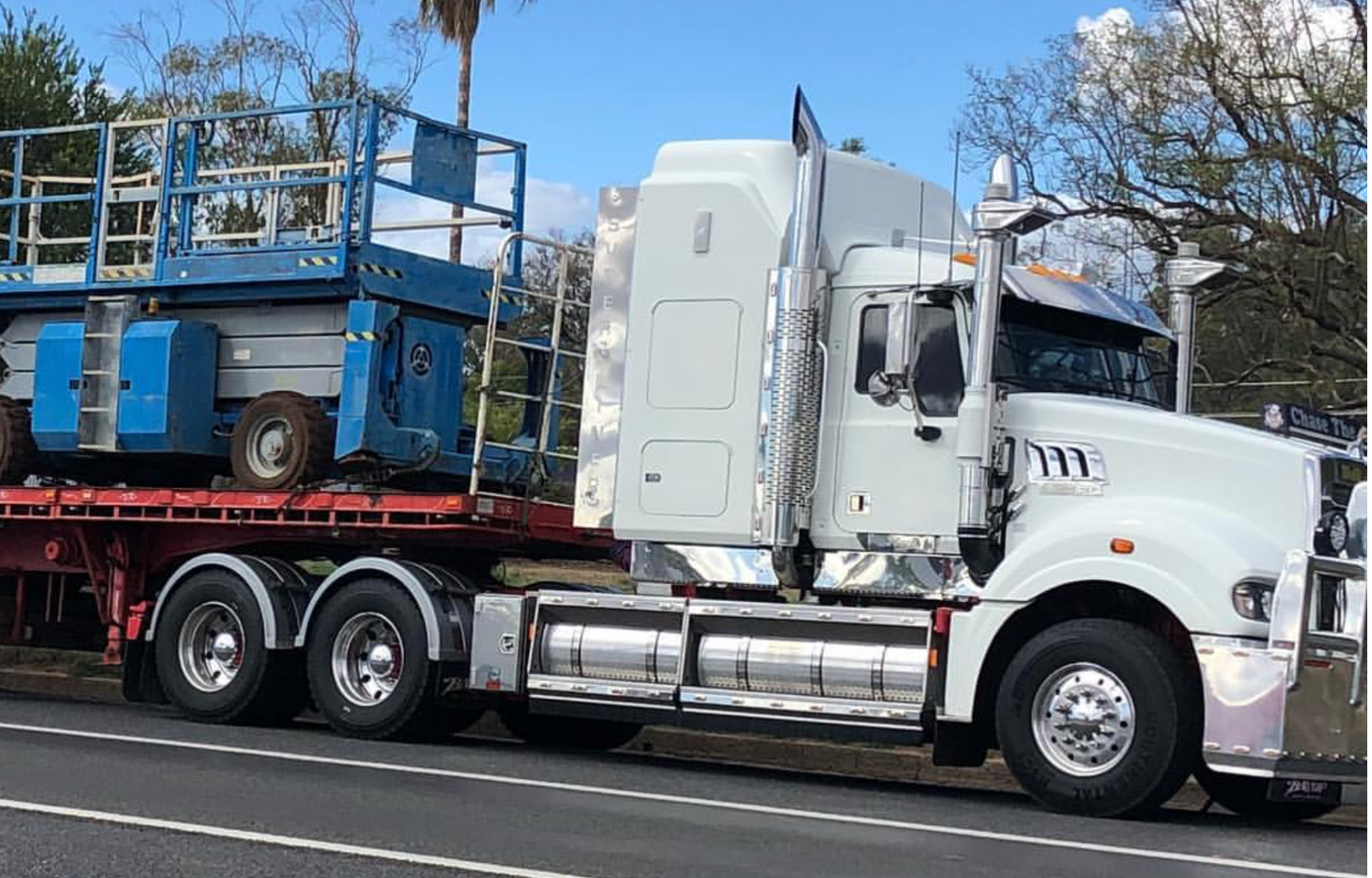 White semi-truck pulling a flatbed trailer with a blue scissor lift on a road — Chewies Tipper Hire & Haulage in Gracemere, QLD