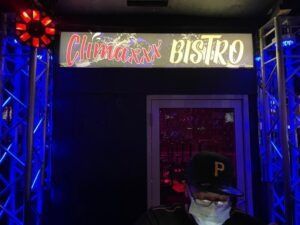 Entrance to Climaxx Bistro with neon sign; DJ wearing a mask and hat.