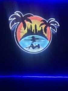 Neon sign with palm trees, cityscape, woman reclining on water; orange, blue, black colors.