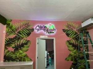 Pink wall with tropical leaf mural and neon sign above an open door; a ladder is nearby.