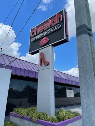 Sign for Cheetah Gentlemen's Club: black, red, and white sign with a shoe and lips, in front of a building with purple trim.
