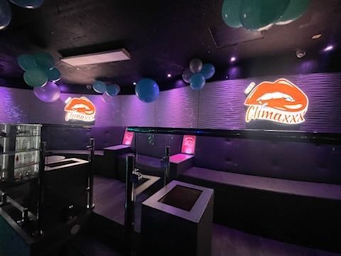 Interior of a nightclub with purple and blue lighting. Booths, balloons, and a lit lip logo are visible.