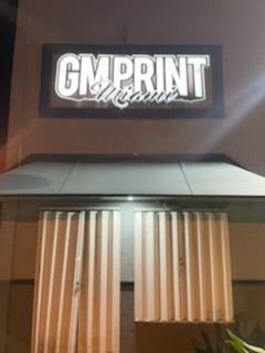 Sign for GM Print Miami above a building's entrance with closed, vertical blinds.