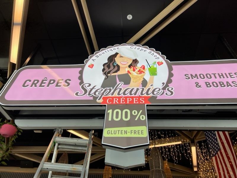 Stephanie's Crêpes sign with a smiling woman, pink background, and gluten-free label.