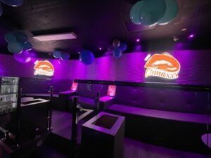 Interior of a club with purple lighting, black booths, and balloons. Logos are displayed.