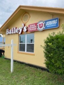 Bailey's scrubs shop with yellow exterior, signage, and a window. A bush and a signpost are in front.