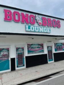Bong Bros Lounge sign in pink and turquoise on a white building. Several windows feature colorful displays.