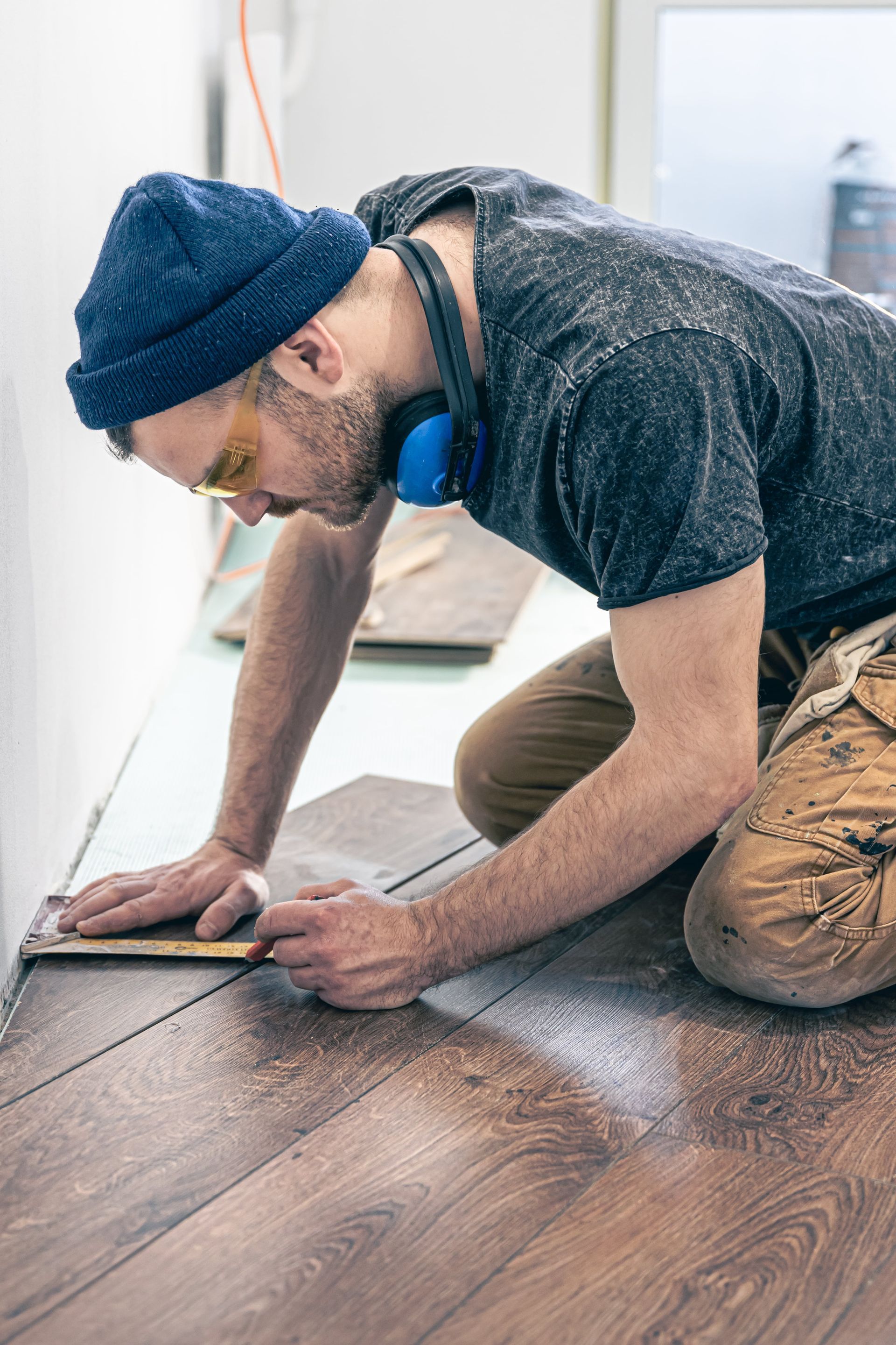 A man is kneeling on the floor measuring a piece of wood.