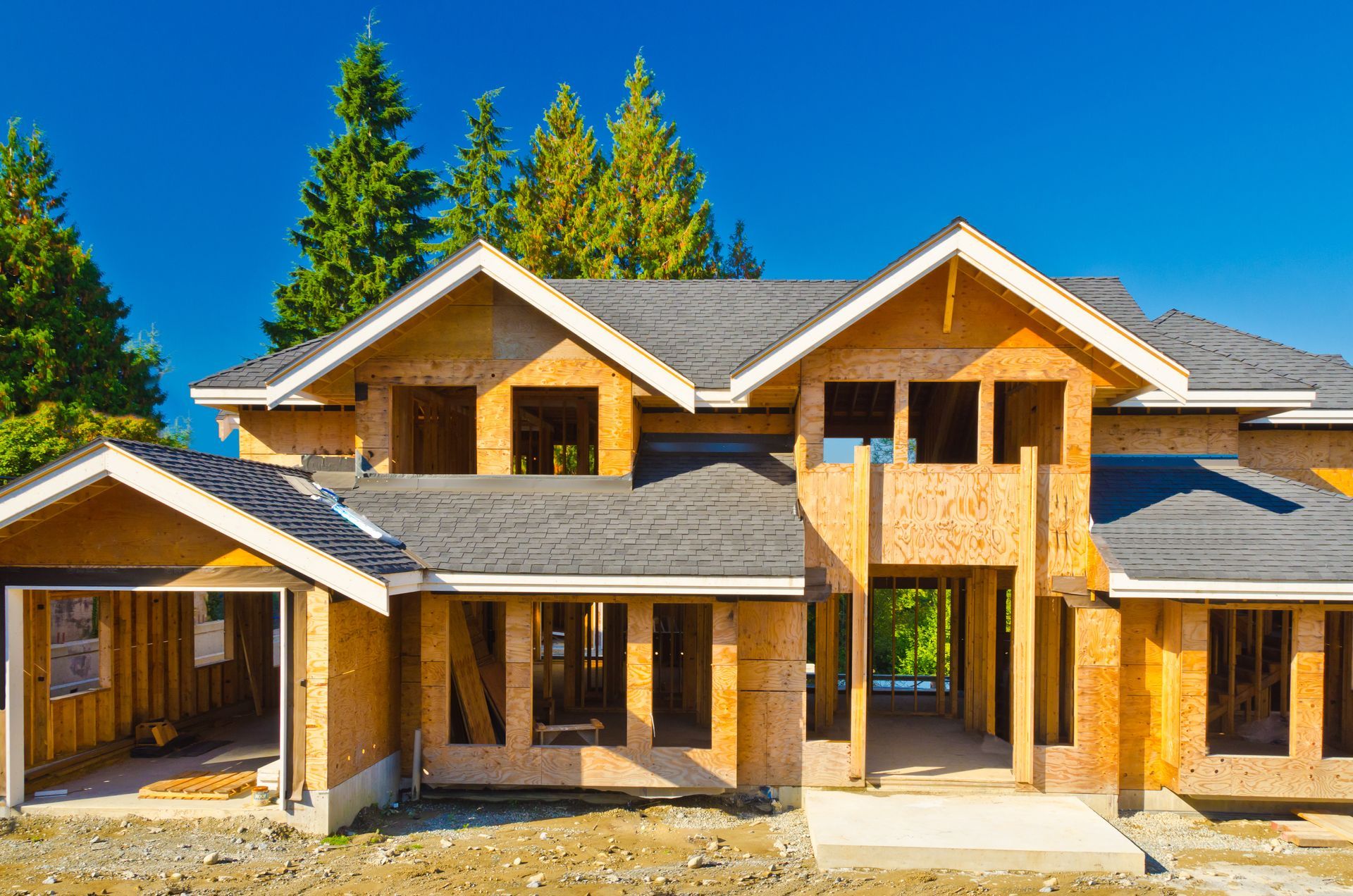 House under construction with wood framing and dark shingles, against a blue sky and trees.