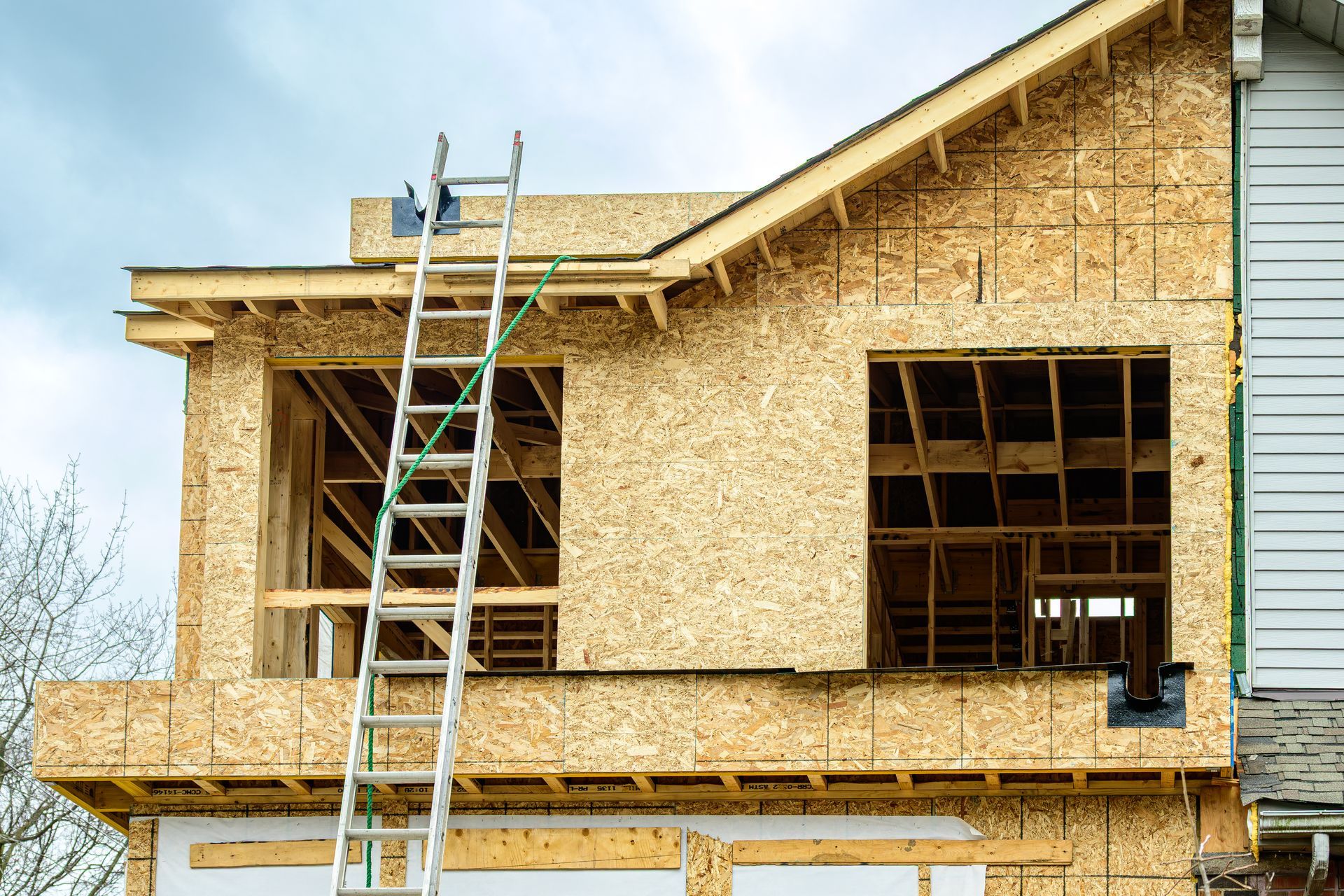 Maison en construction avec charpente apparente, fenêtres et échelle appuyée contre le côté. Ciel couvert.