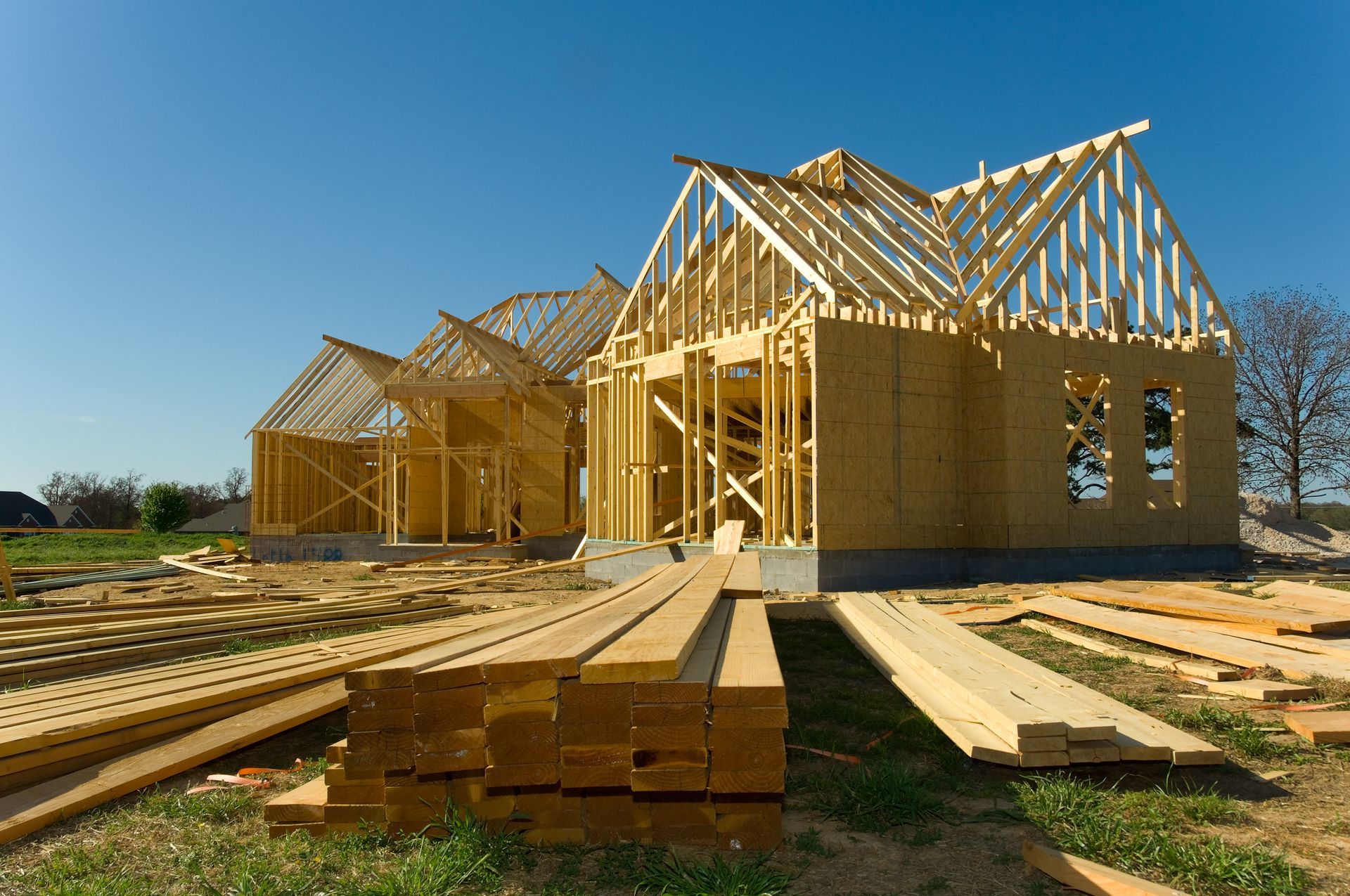 Construction de plusieurs maisons en bois, avec charpente apparente et piles de bois, par une journée ensoleillée.