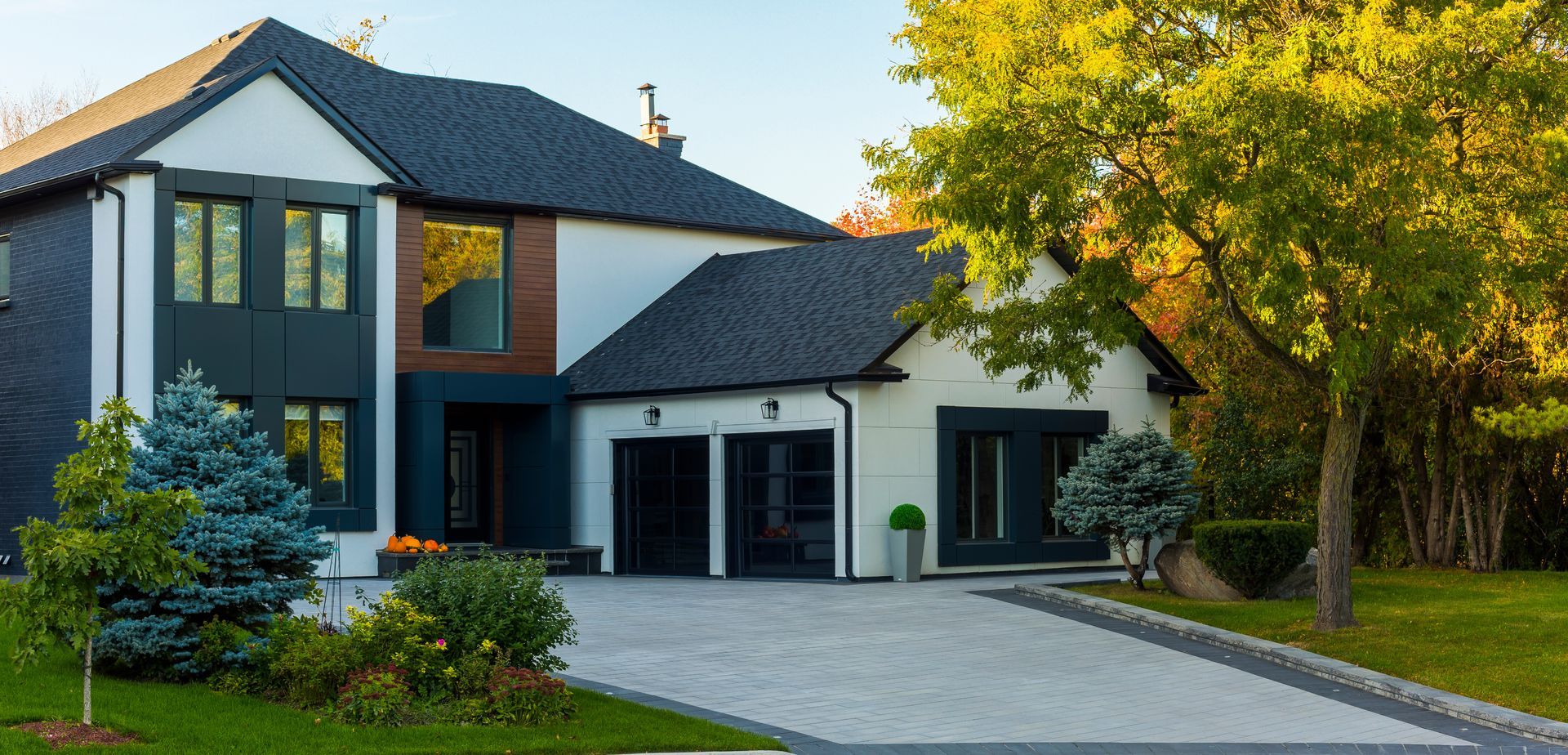 Modern two-story house with black roof and garage doors, white siding, and a long paved driveway. There are trees and shrubs in the front yard.