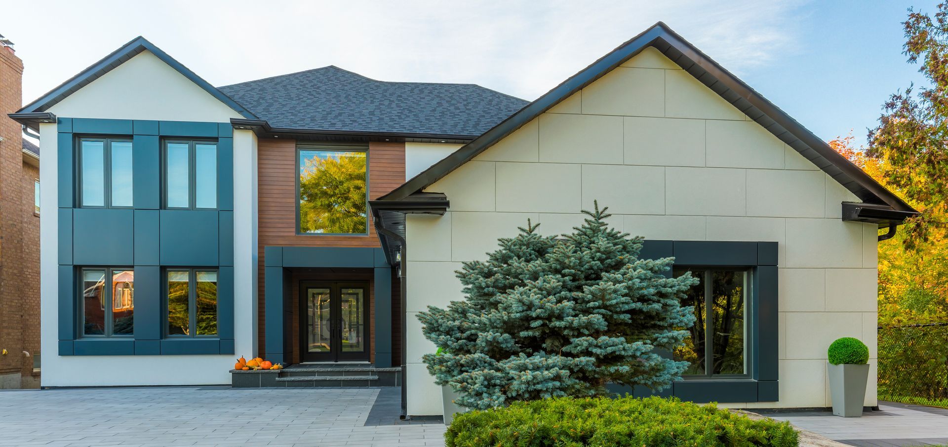Modern two-story house with a blue-gray facade, dark roof, and a light-colored, paneled garage. A blue spruce and green hedges frame the entrance.
