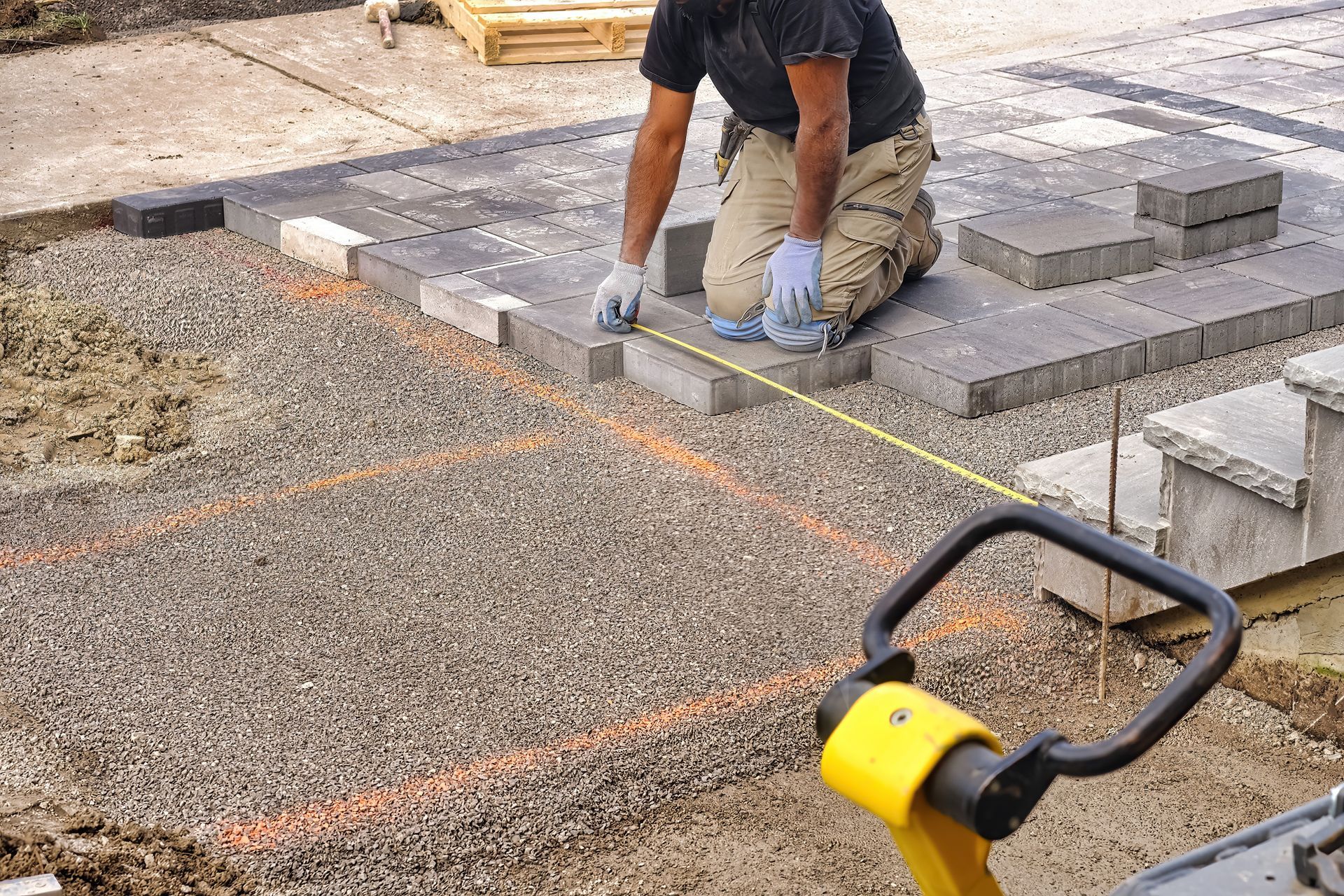 Person laying paving stones, using a tape measure, on a gravel base. Construction site with a compactor nearby.