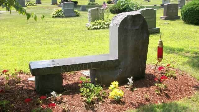 A stone bench-style headstone in a cemetery with flowers, grass, and other headstones.