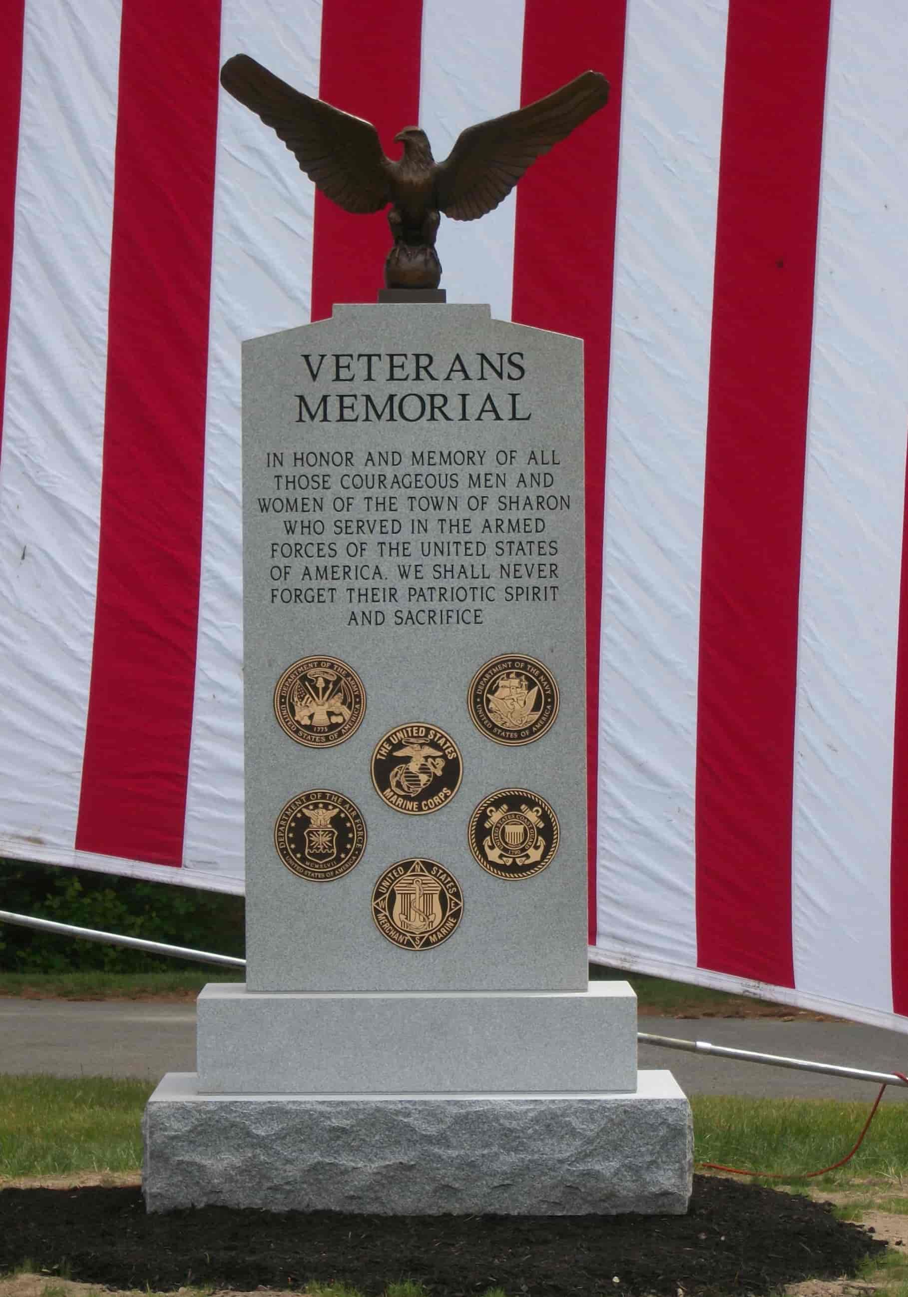 Veterans Memorial with bronze eagle atop. Gray stone with dedication, service emblems, and American flag background.