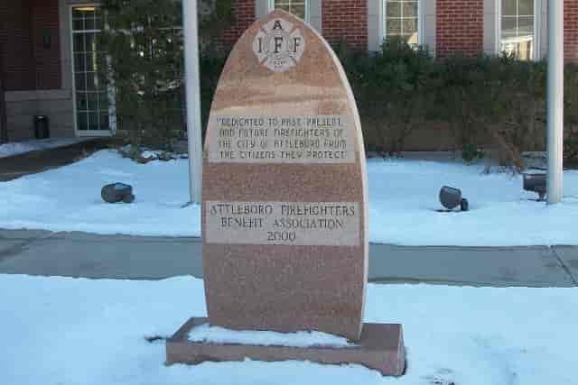 A granite memorial for firefighters with text, snow on the ground, and building in the background.