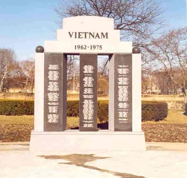 Vietnam War memorial with names inscribed on black columns. White stone structure, grass, trees.