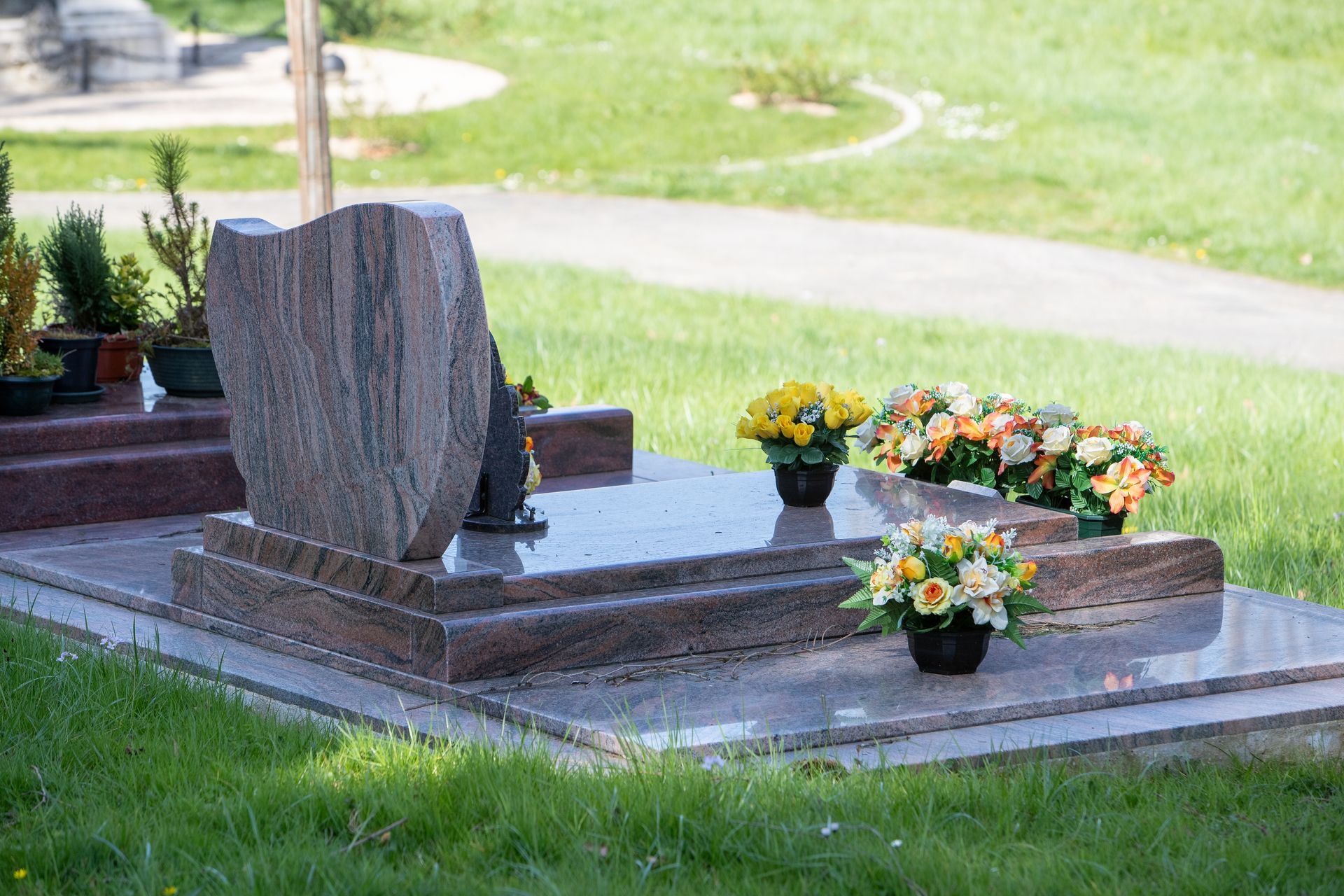 Grave with a pinkish granite headstone and base; flowers in pots sit around it, set in a grassy cemetery.