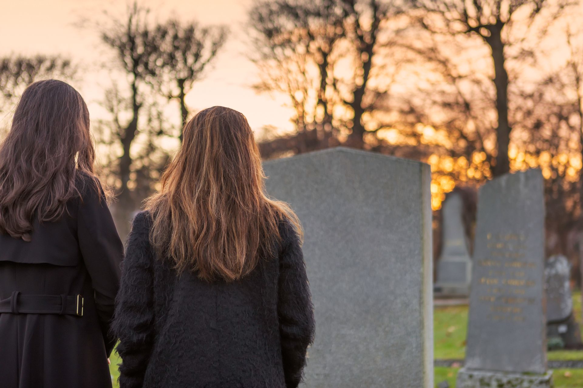 Two women in black coats at a cemetery, looking at headstones during a sunset.
