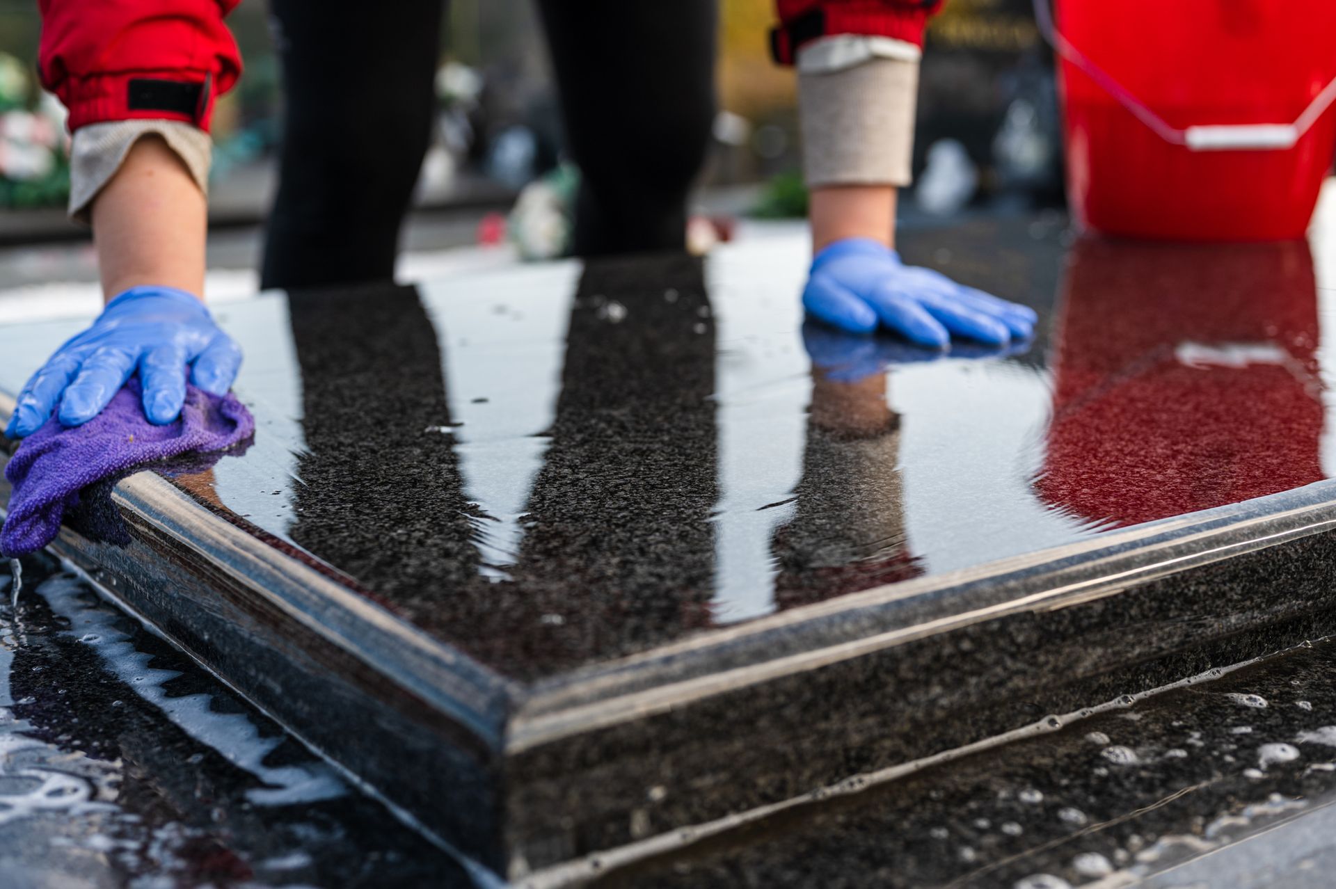 Hands in blue gloves wiping a wet, shiny black surface with a purple cloth; red bucket visible.
