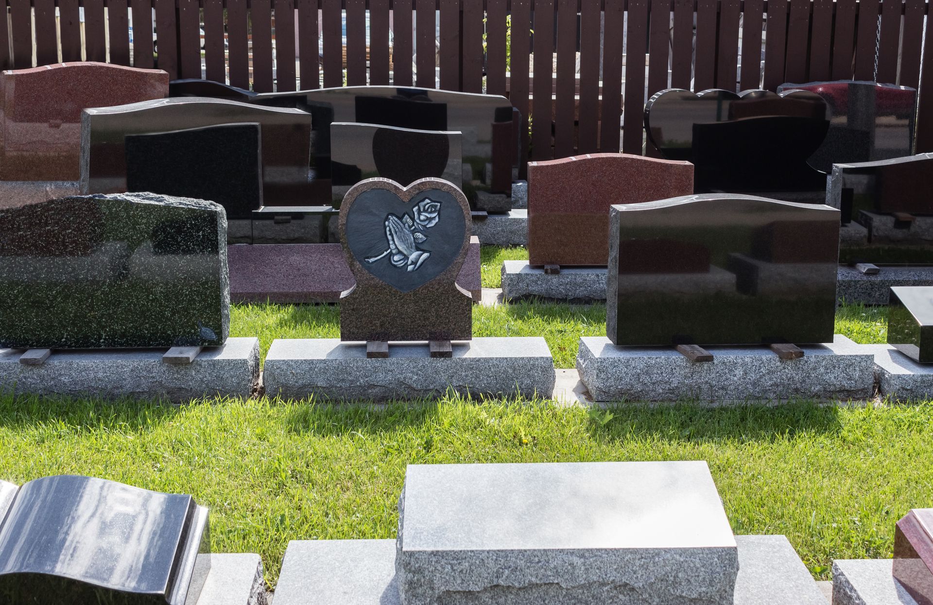 Headstones on display, various shapes and colors, on a grassy surface. Brown wooden fence background.