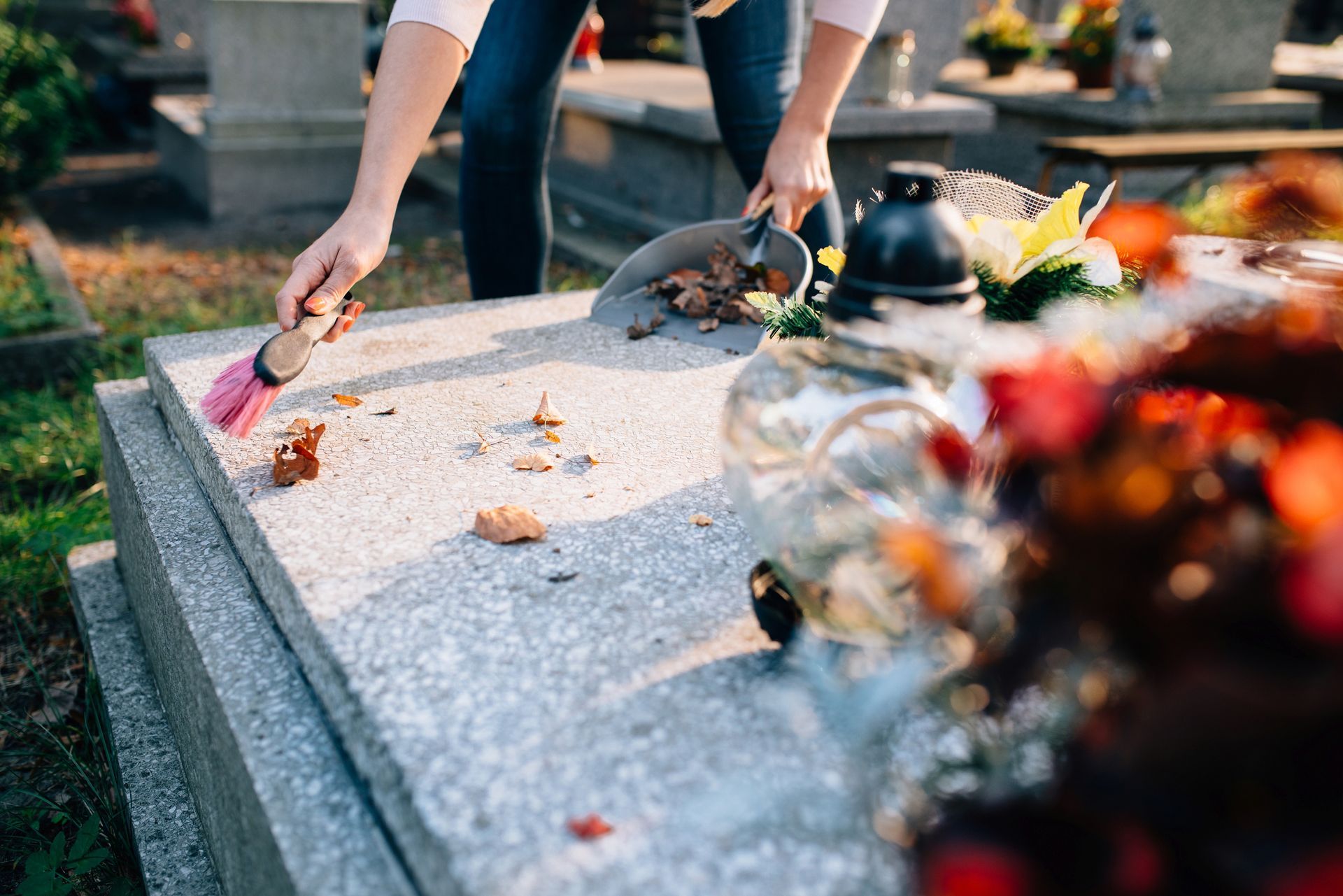 Person cleaning a tombstone with a brush and scoop in a sunlit cemetery.