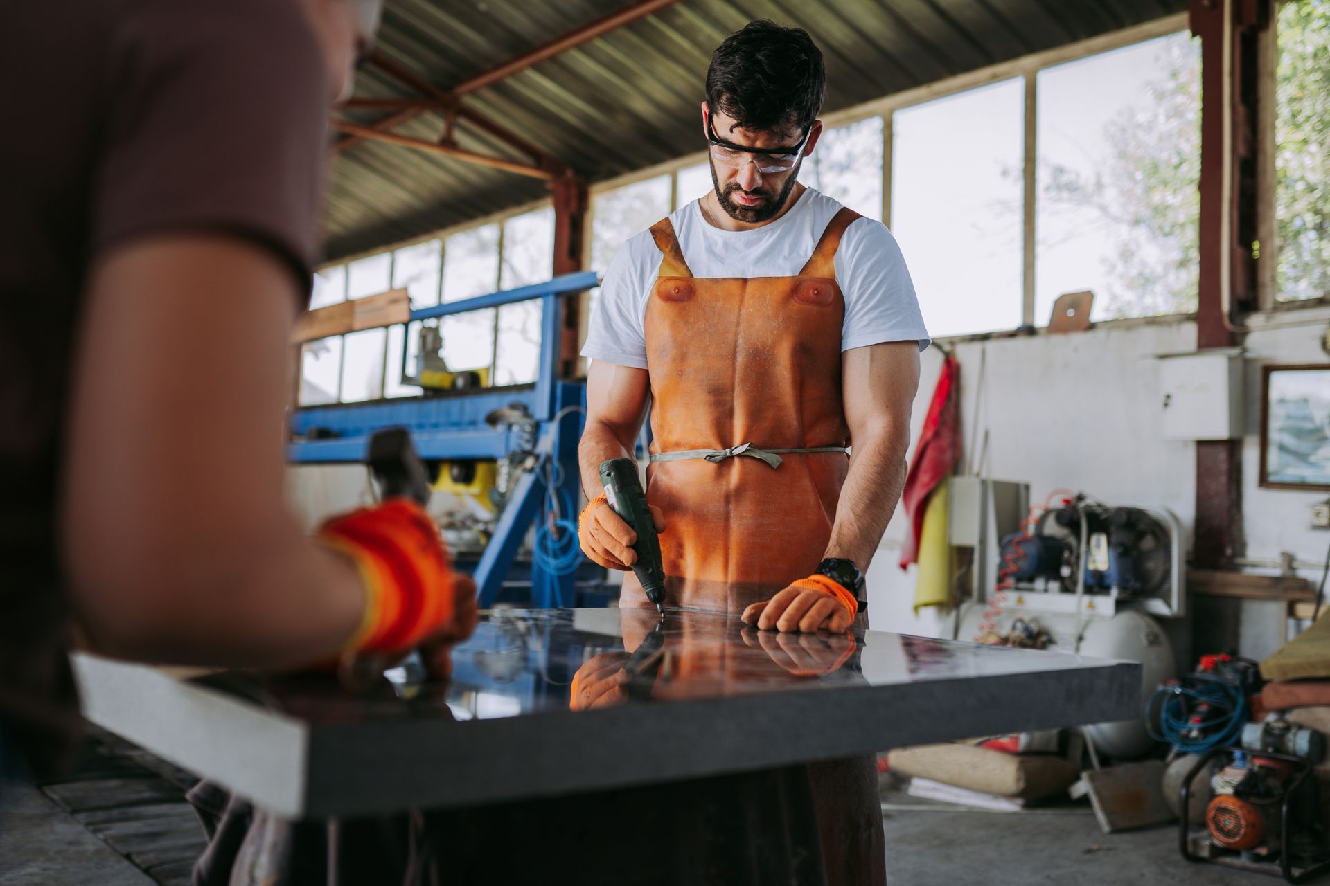 Two men working on a dark, shiny stone surface in a workshop. One man uses a power tool, wearing safety glasses, apron, and gloves.