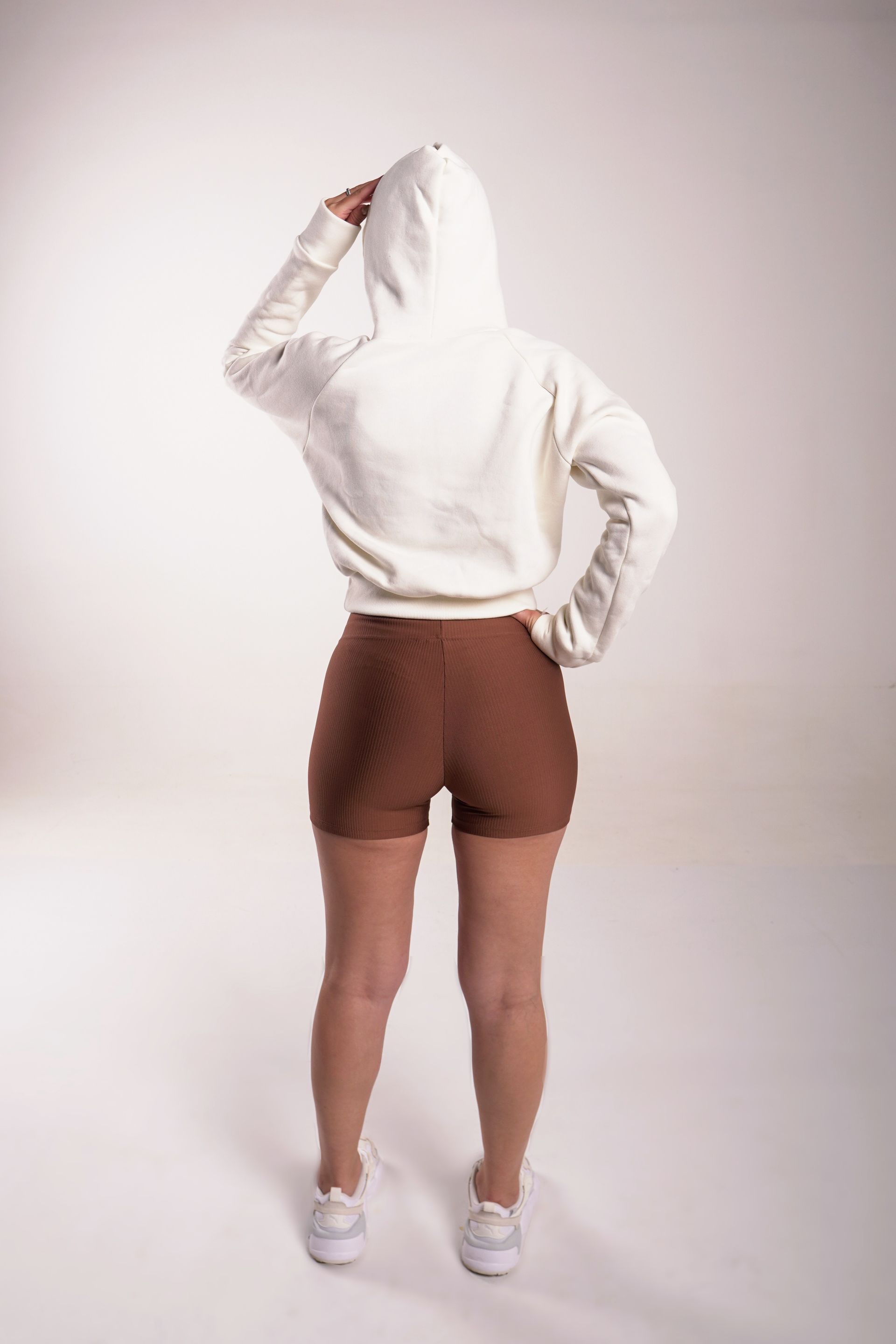 Mujer con sudadera con capucha beige y pantalones cortos marrones, de pie con la mano en la cabeza, de espaldas, fondo blanco.