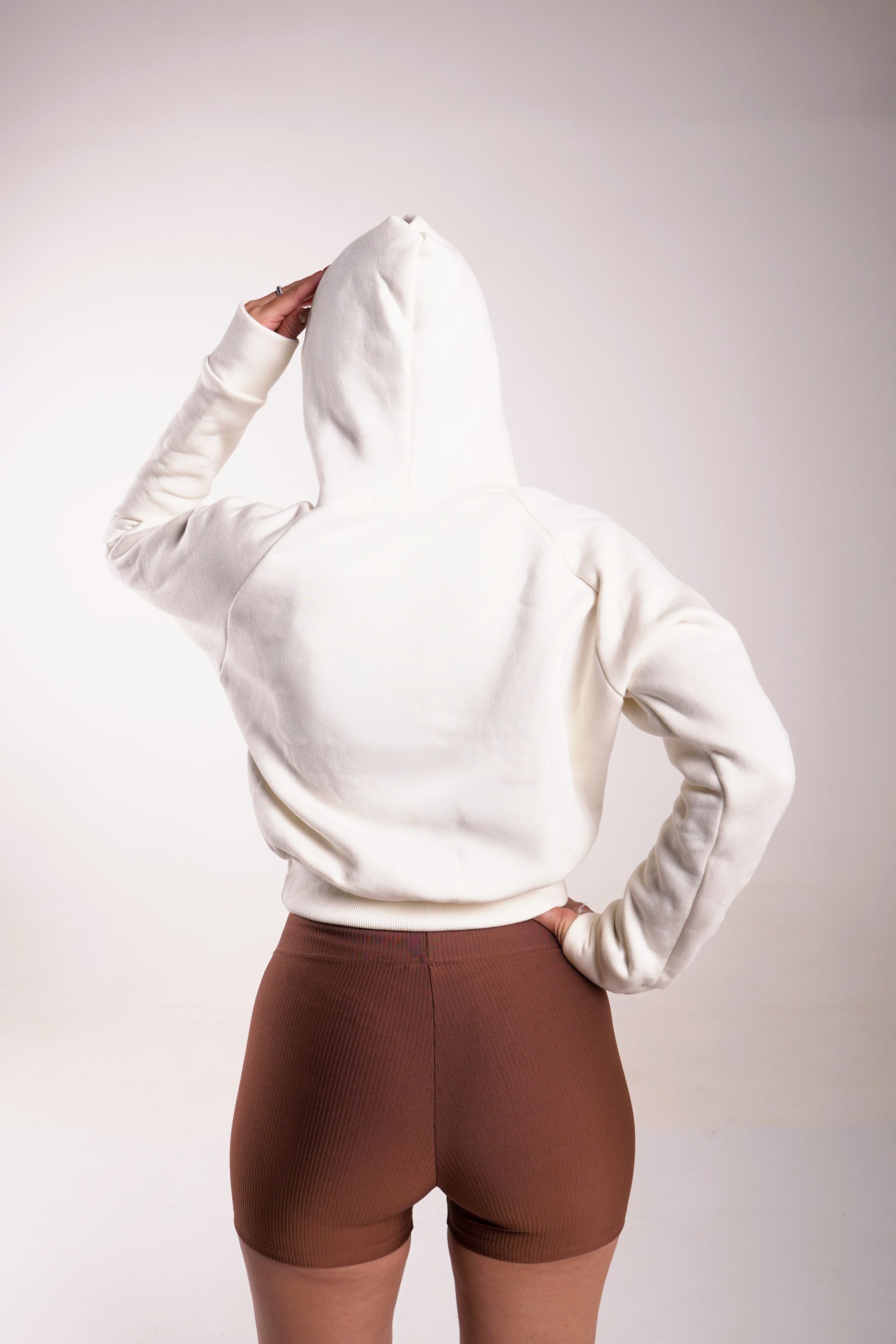 Mujer con sudadera con capucha beige y pantalones cortos marrones, de pie con la mano en la cabeza, de espaldas, fondo blanco.