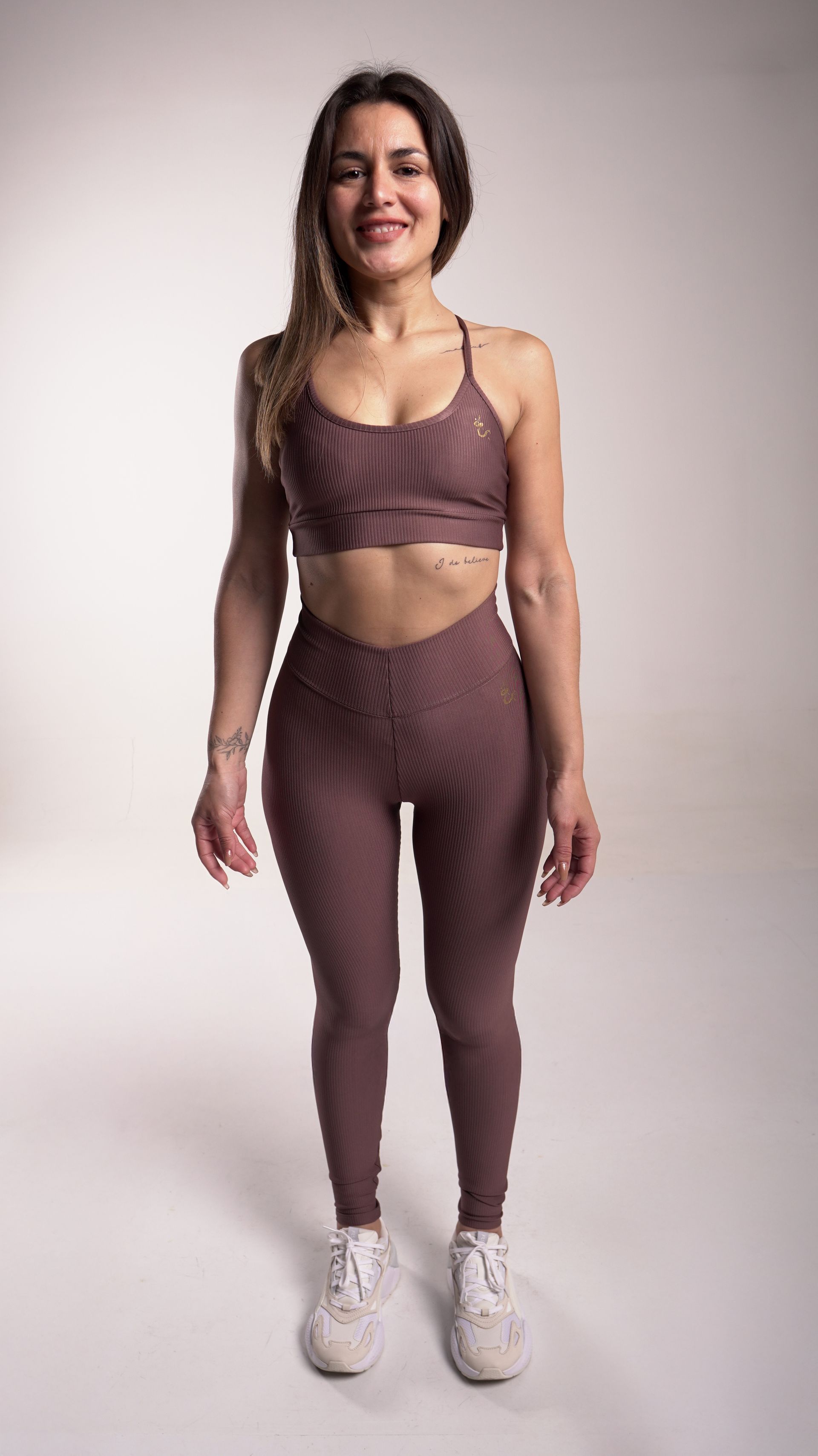 Mujer con un conjunto deportivo de color malva, sonriendo, de pie frente a un fondo blanco.