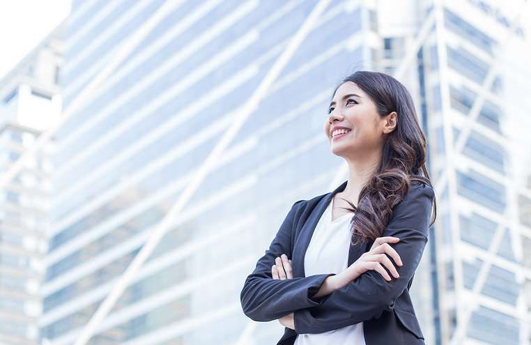 A woman in a suit is standing in front of a tall building with her arms crossed.