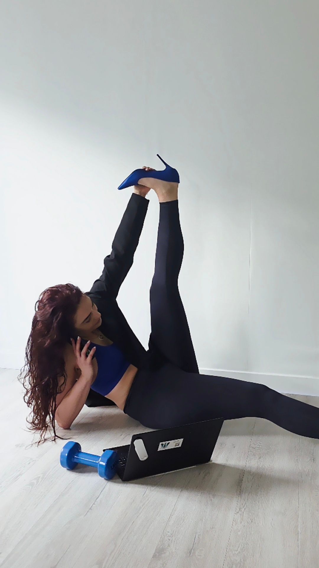 Een persoon in een blazer, crop top en legging houdt één blauwe schoen met hoge hak vast terwijl ze op de grond aan de telefoon praat.