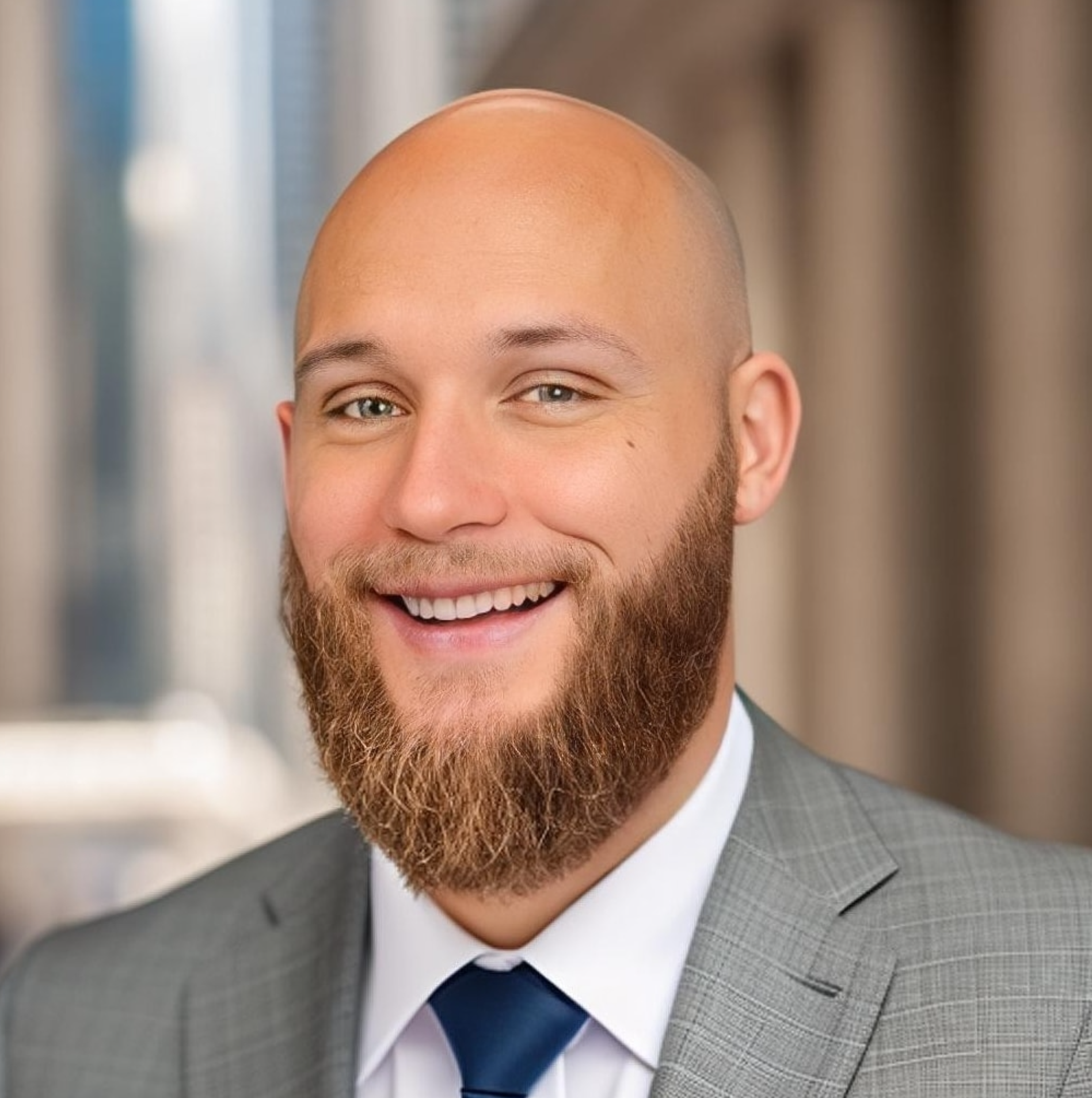 Nick Byrne with a full beard, wearing a gray suit and navy tie, smiling in front of a blurred urban background.