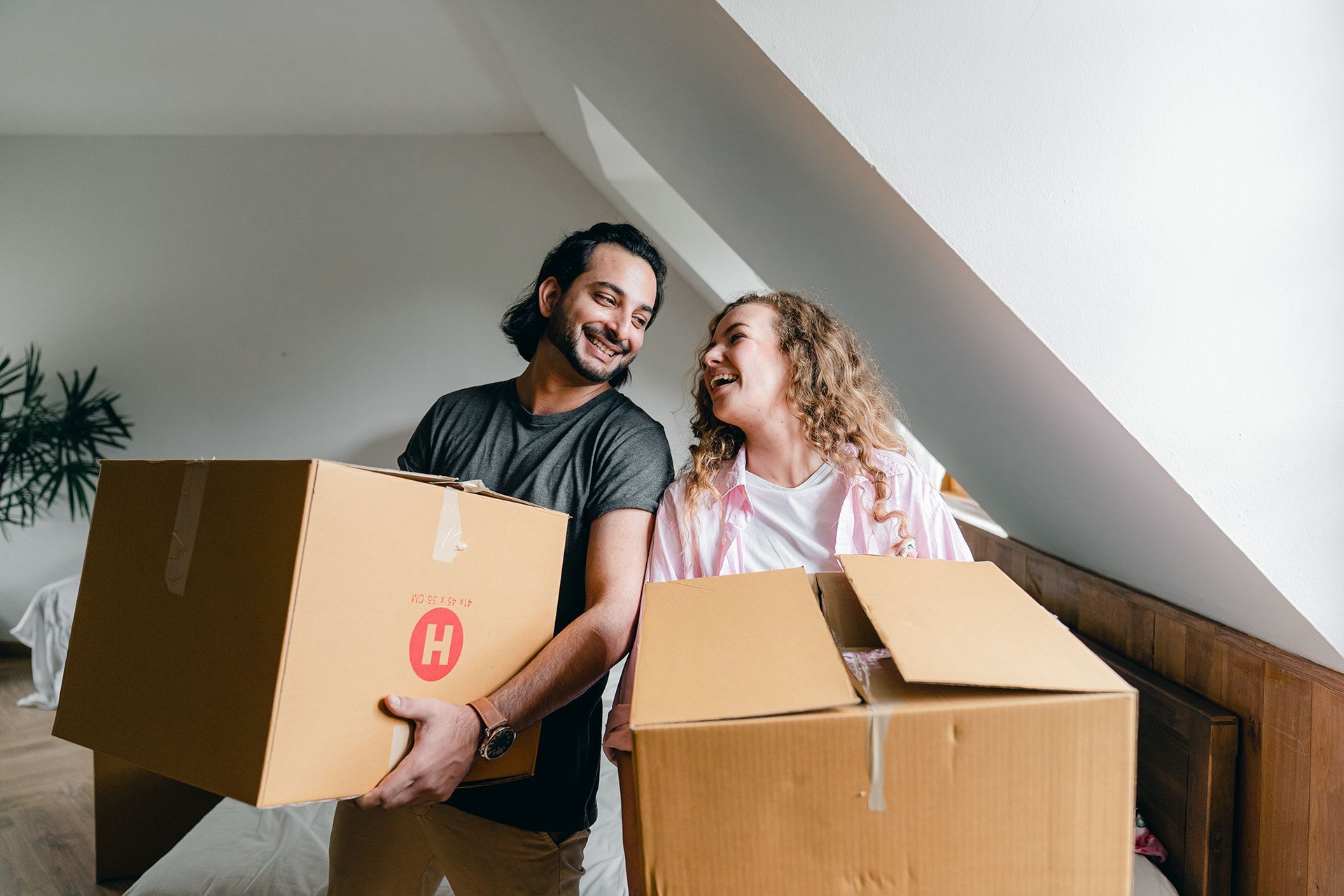 Couple carrying cardboard moving boxes, smiling, inside a home.