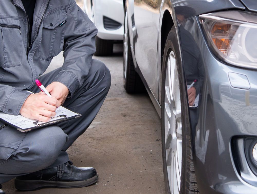 Mechanic inspecting car tire, writing on clipboard, wearing gray work suit.