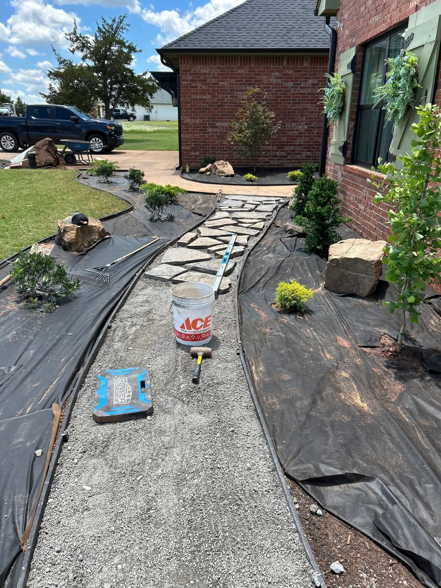 Gravel garden path with black landscape fabric, small plants, and books near a brick house.