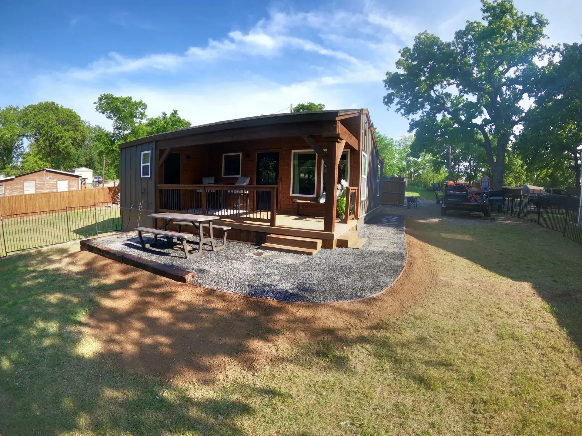 Small wooden cabin with porch and gravel patio in a sunny backyard, surrounded by trees and grass.