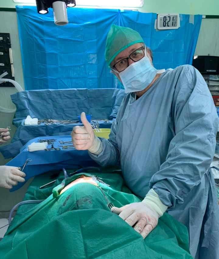 Un chirurgo, con indosso camice, mascherina e cuffia, fa il segno del pollice in su mentre esegue un intervento chirurgico in sala operatoria.