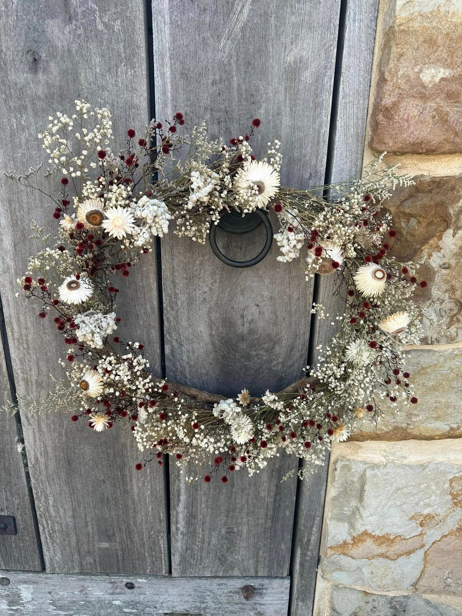 Wreath made of dried flowers and berries hanging on a gray wooden door with a stone wall.