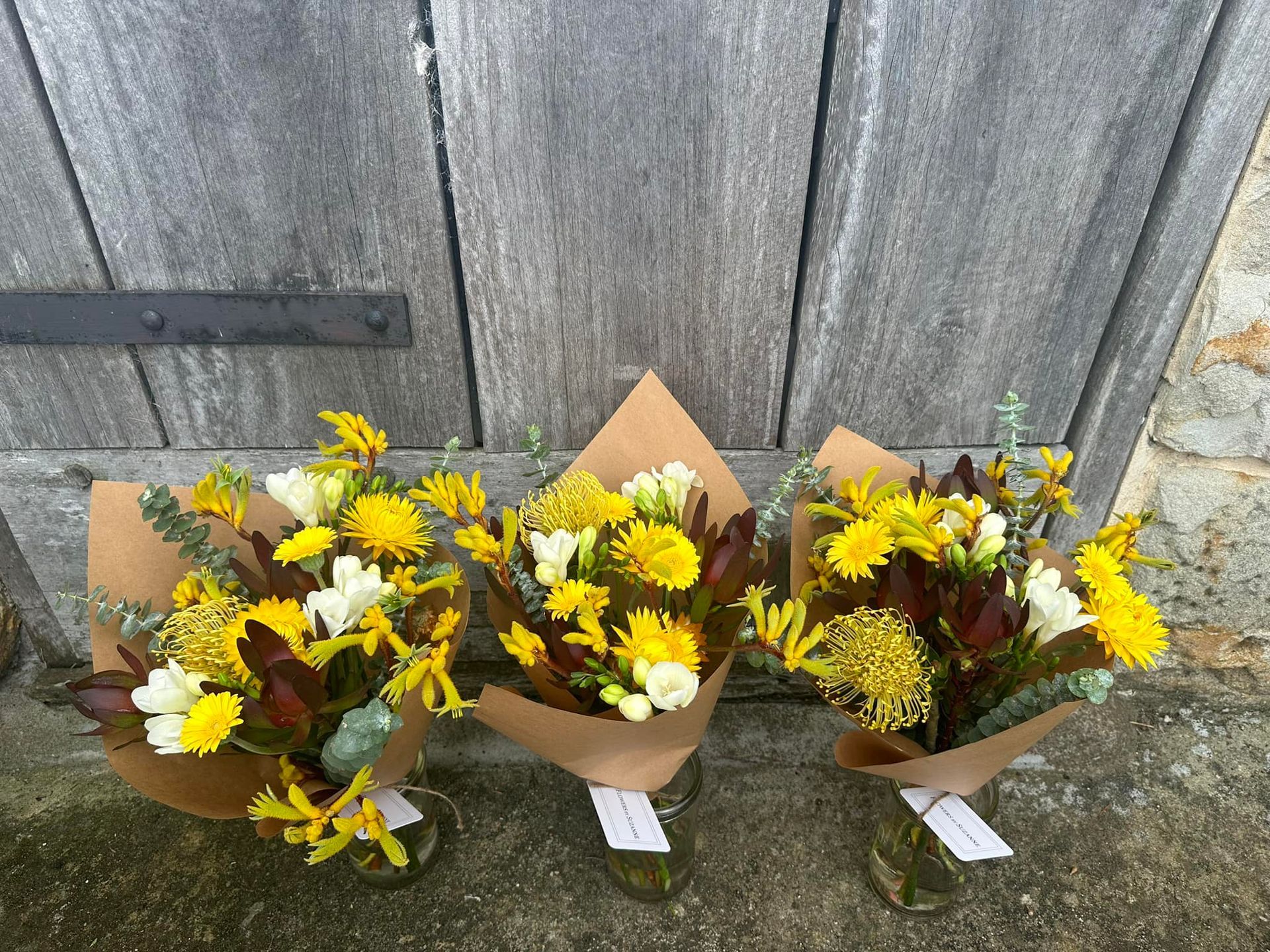 Three yellow and white flower bouquets wrapped in brown paper, sitting in glass jars.