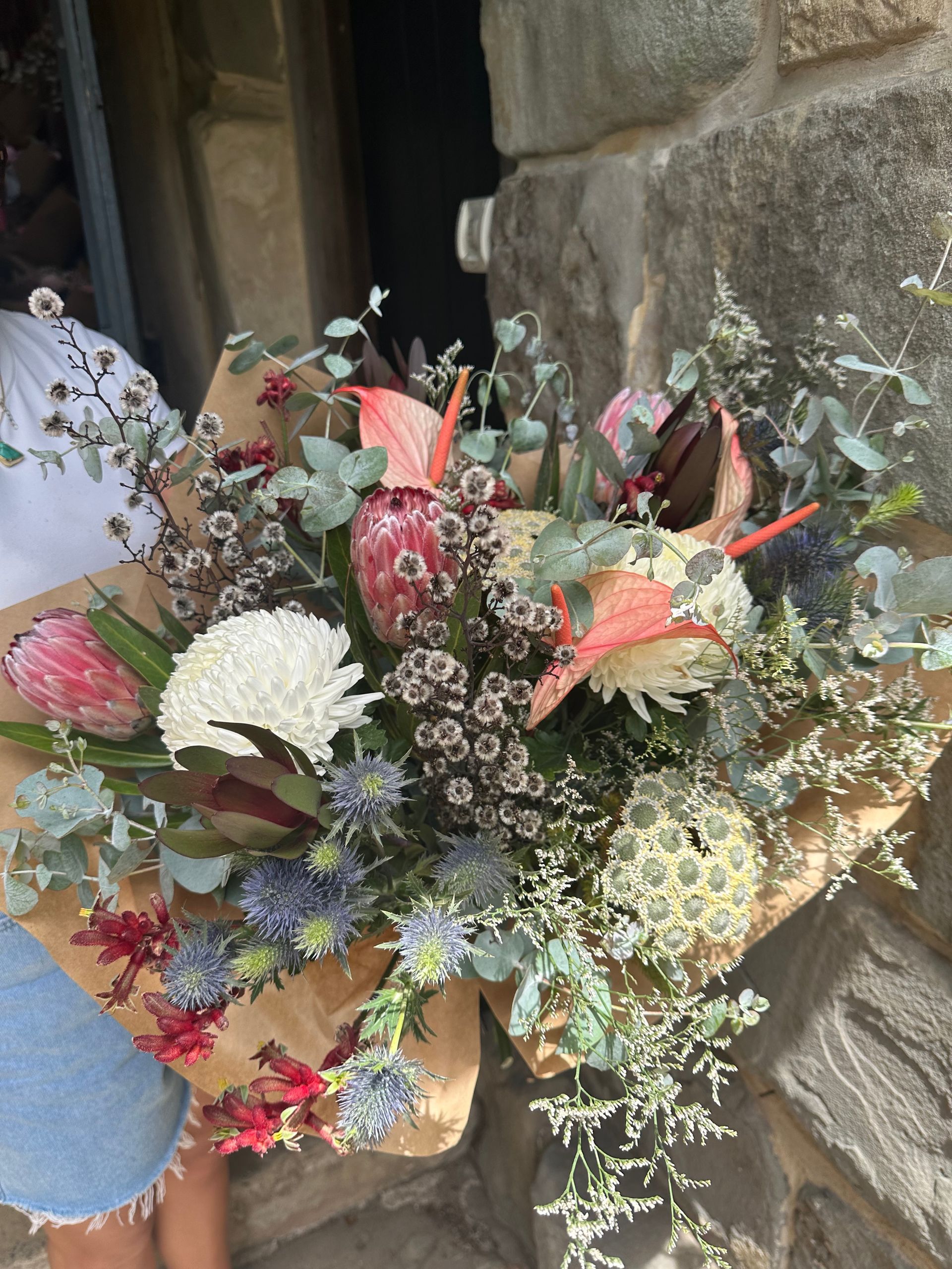 Person holding a bouquet of colorful flowers wrapped in brown paper, outside a stone building.