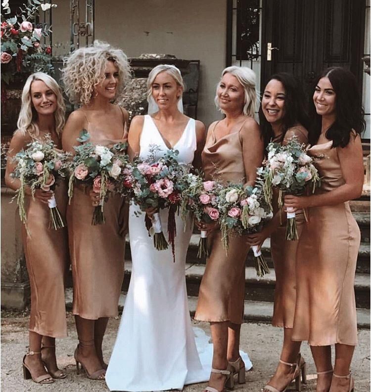 Bride in white gown with bridesmaids in gold dresses, holding bouquets, standing outdoors.