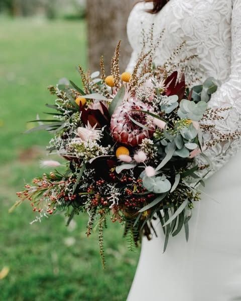 Woman in white dress holds a rustic bouquet with protea, eucalyptus, and wildflowers.