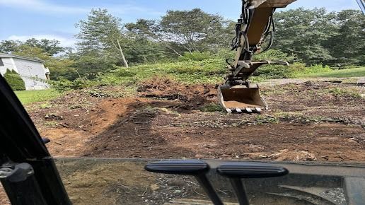 Excavator digging in dirt, viewed from inside cab, with trees and a building in the background.