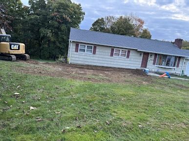 A yellow CAT excavator works on the front lawn of a beige house.