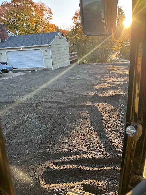 View from excavator cab, ground prepared for paving. Sunlit driveway in front of a garage; trees with fall foliage in background.