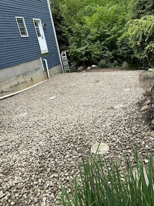 A gravel yard beside a blue house, with a ladder and greenery in the background.