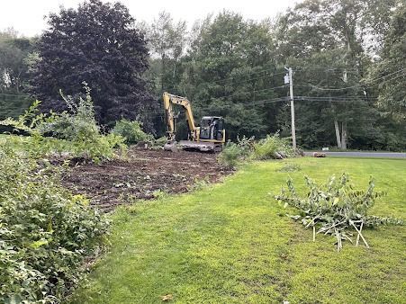 A yellow excavator clearing a muddy patch of land next to a green lawn with trees in the background.
